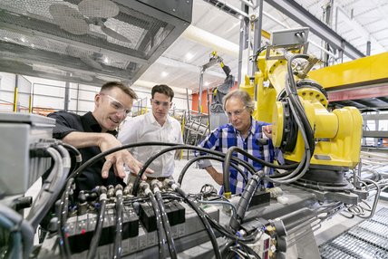 Trois hommes avec lunettes de protection discutent autour d'une machine avec de nombreux câbles dans un atelier