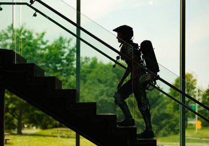 Une étudiante équipe d'un exosquelette gravit un escalier devant un mur vitré donnant vue sur la nature.