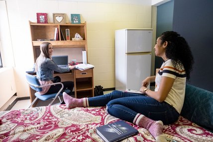 Dans une champbre de résidence, deux étudiantes, l'une assise au bureau et l'autre sur le lit, discutent.