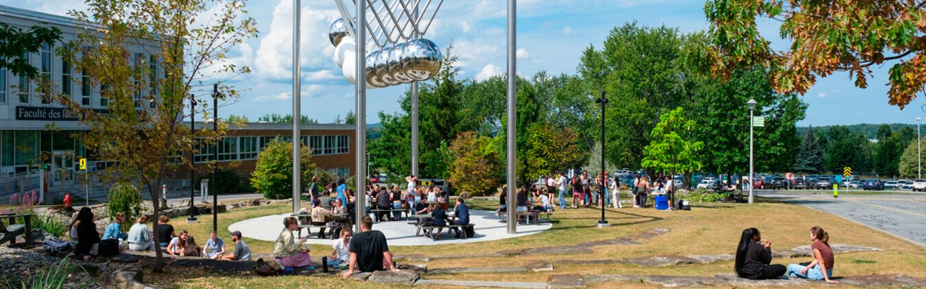 Plusieurs étudiantes et étudiants assis au soleil à proximité de l'oeuvre magistrale au coeur du campus principal.